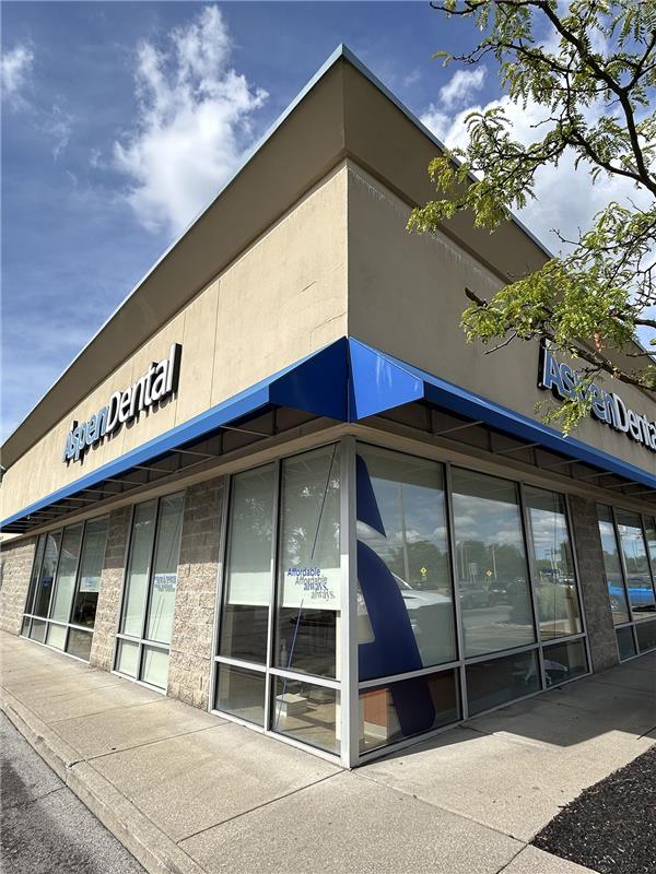 Exterior view of an Aspen Dental facility with large glass windows, a blue awning, and the company's white logo mounted on the light-colored facade.