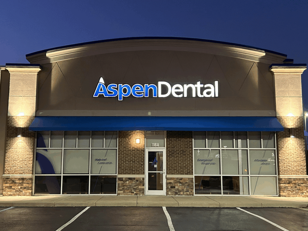 Exterior night view of an Aspen Dental office with the logo lit up in white and blue, a blue awning over the windows, and interior lights illuminating the space, showing a well-lit parking lot in the foreground.