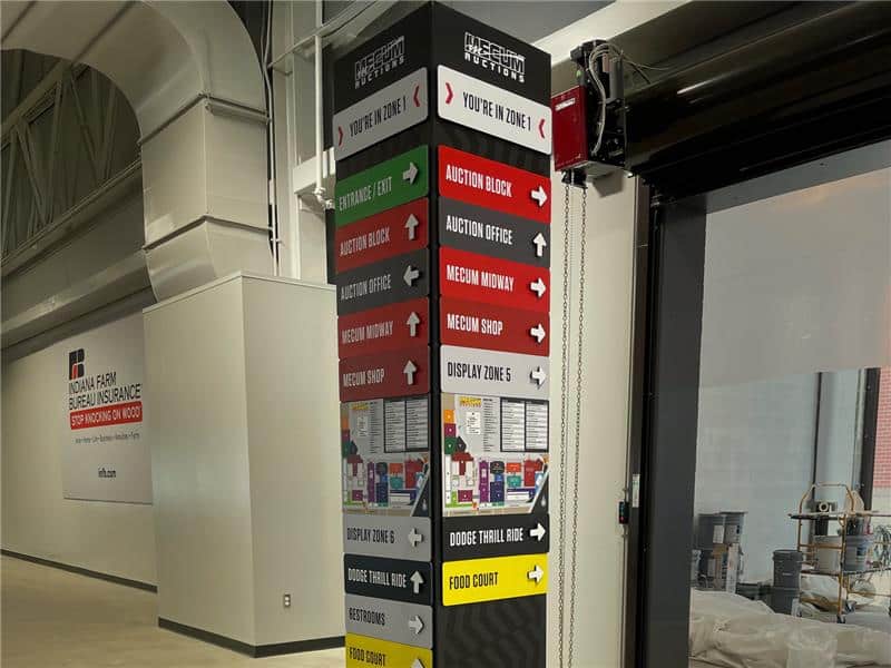 A tall, multi-sided interior signpost with stacked, colored directional arrows pointing to various Mecum Auction areas, including "Registration," "The Auction Block," "Registration Shop," and "Restrooms."