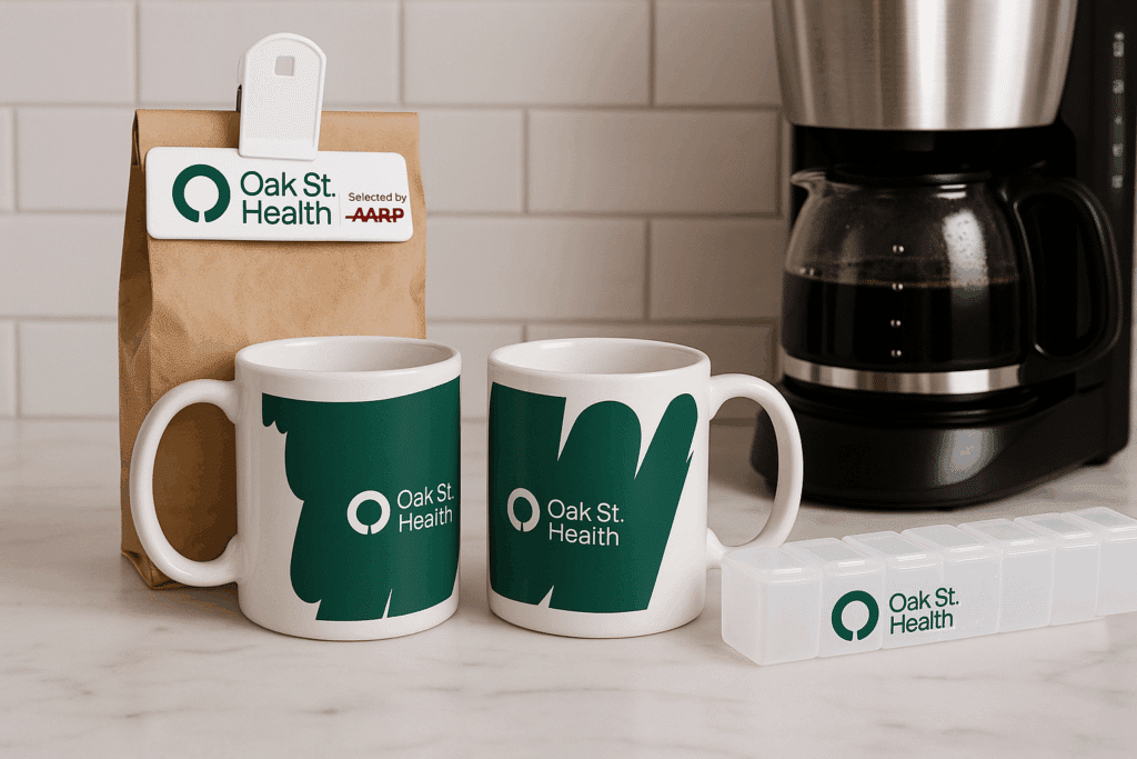 Two white ceramic coffee mugs and a rectangular white pill organizer, all custom branded with the dark green and white "Oak St. Health" logo, sit next to a paper bag with the "Oak St. Health + AARP" logo.