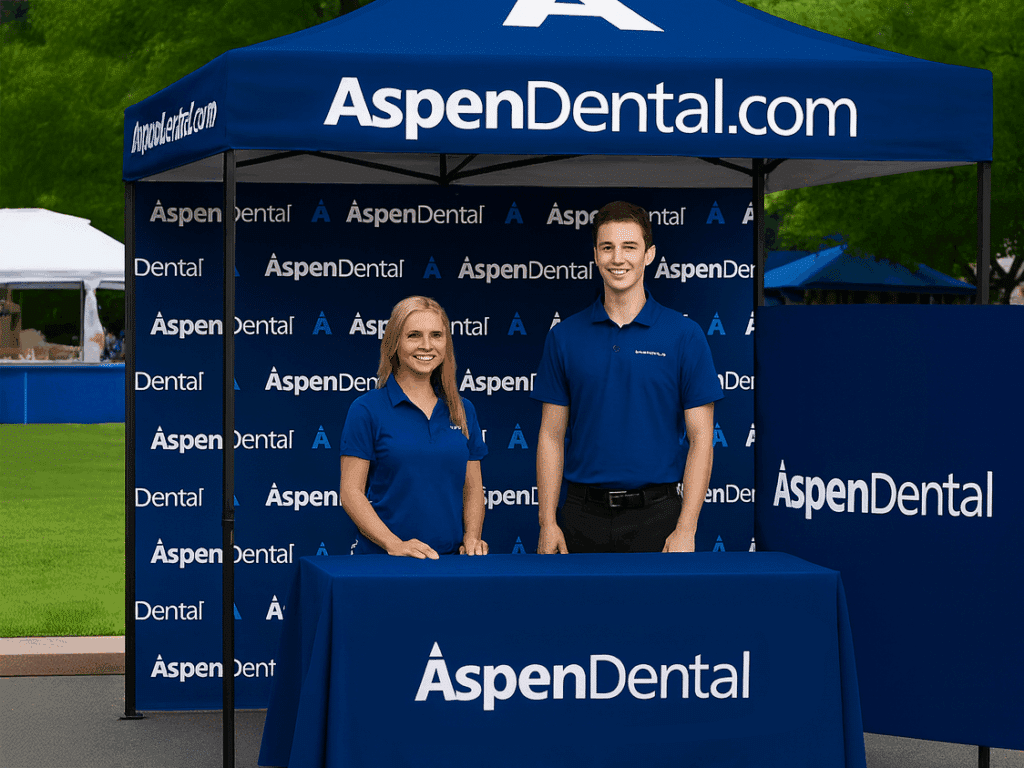 Two smiling Aspen Dental employees, a woman and a man, stand behind a covered table in front of a branded dark blue tent display with the "AspenDental.com" logo.