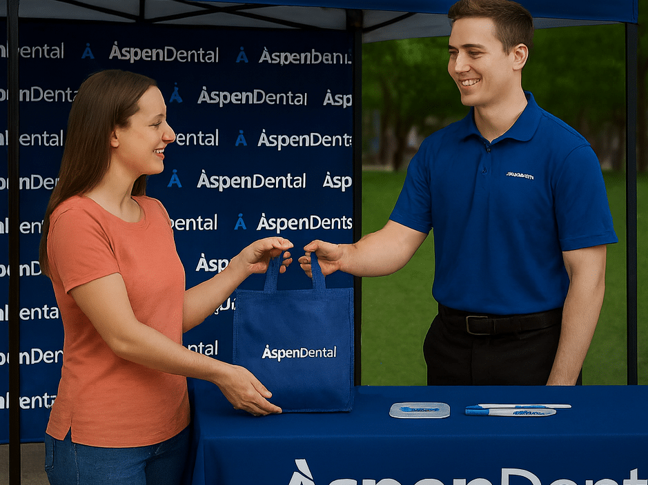 A smiling female Aspen Dental employee in an orange shirt and blue jeans hands a dark blue branded bag to a smiling male employee in a dark blue polo shirt, across a covered table at a promotional event with a large, branded "Aspen Dental" backdrop.