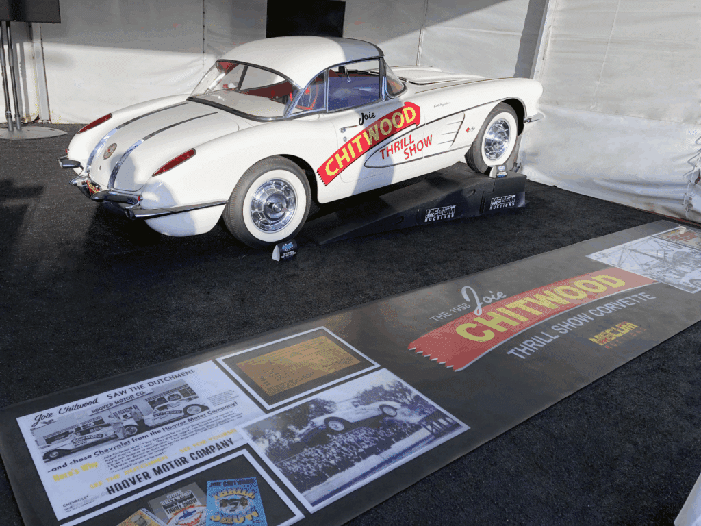 A white 1958 Chevrolet Corvette (C1) is displayed on a ramp in an indoor exhibit space, next to a large floor decal promoting the Joie Chitwood Thrill Show and Mecum Auctions.