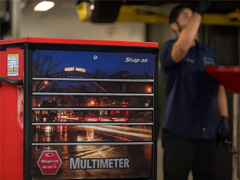Close-up of a red Snap-on tool chest with a printed street scene graphic that includes a "Multimeter" control panel. A technician in a dark blue shirt is partially visible on the right.