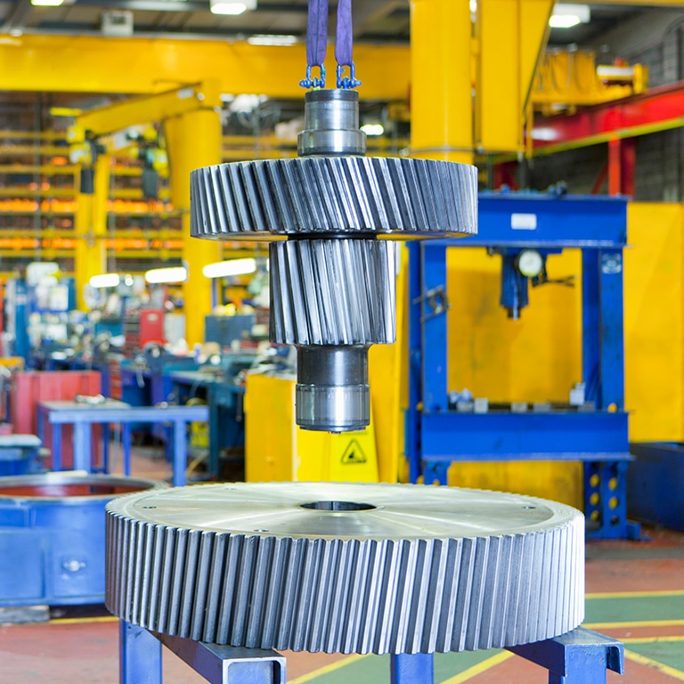 A heavy industrial metallic gear component suspended by blue riggings above a large gear ring in a manufacturing facility.