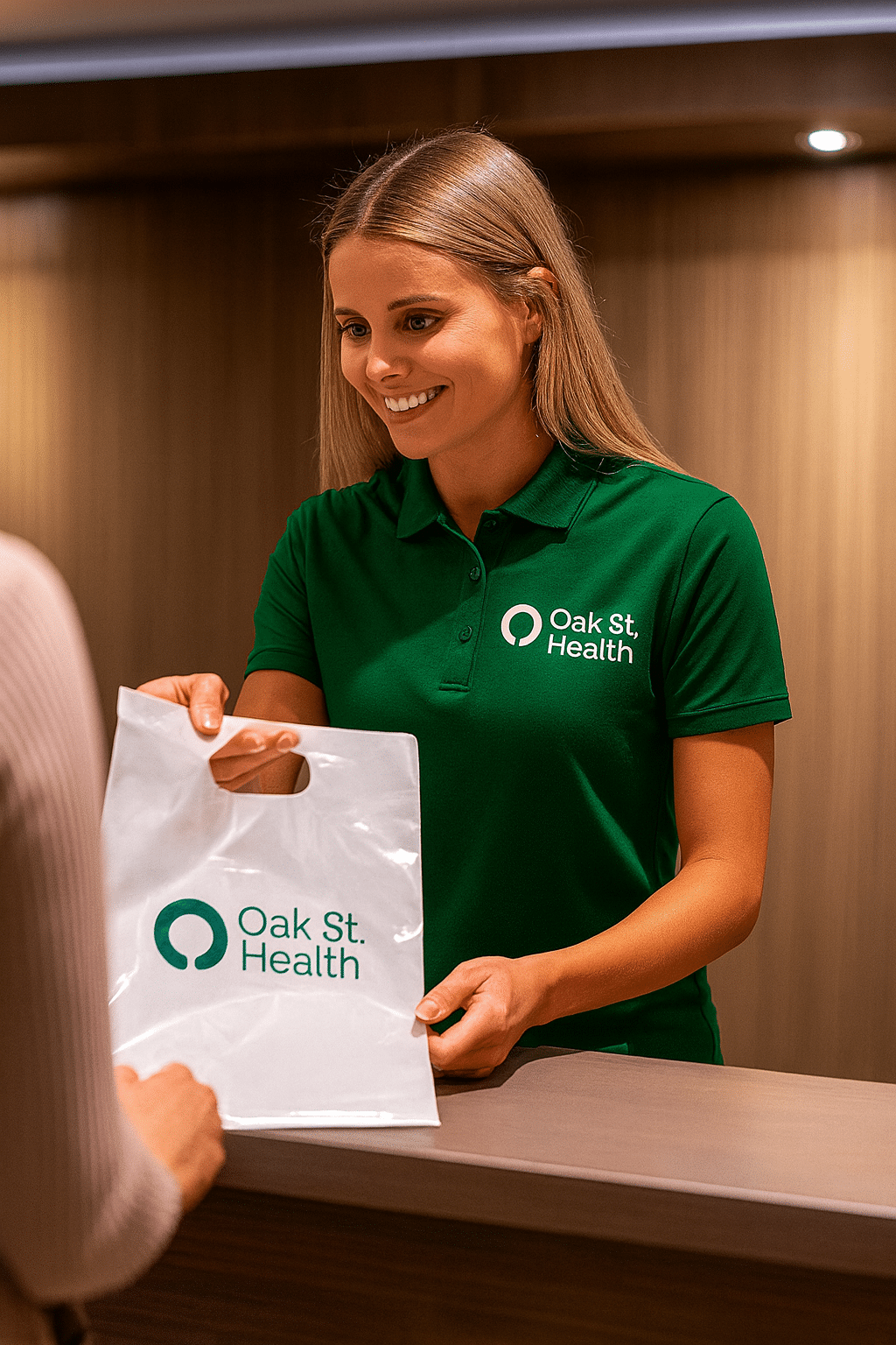 A smiling female employee in a dark green Oak St. Health polo shirt hands a white plastic shopping bag with the Oak St. Health logo to a person across a light-colored counter.