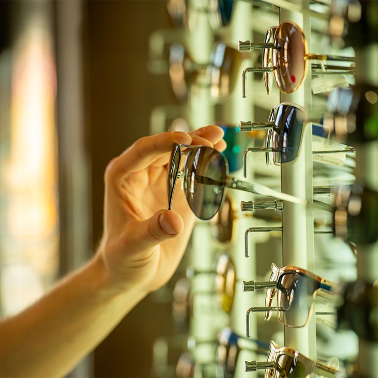 Customer hand selecting fashionable sunglasses from illuminated optical store display.