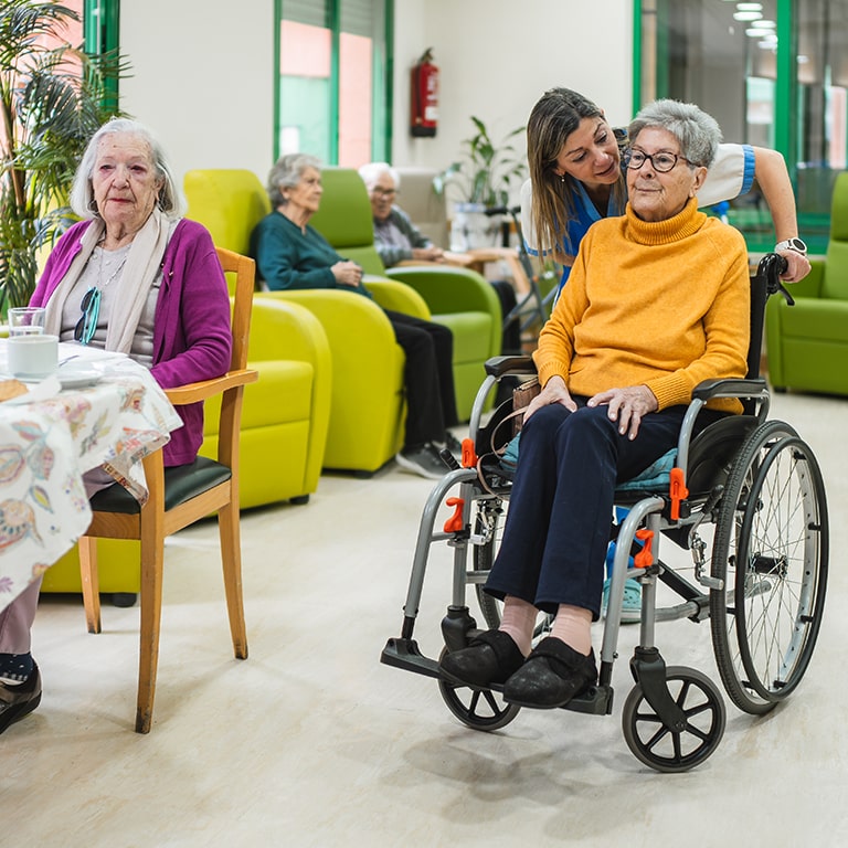 Senior women sitting in wheelchair and at table, a young caregiver assisting in a nursing home or retirement facility